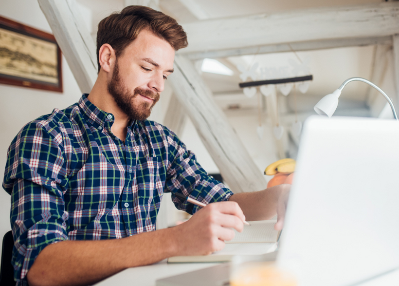 Man sitting down at desk with book and pencil and laptop and desk light in the background.